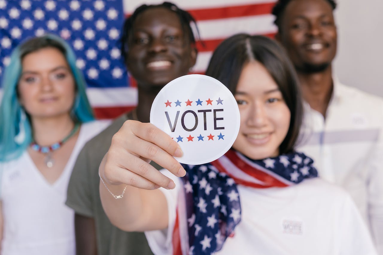 Four young people standing in front of the U.S. flag smiling with one young asian woman holding up a sticker that says vote.